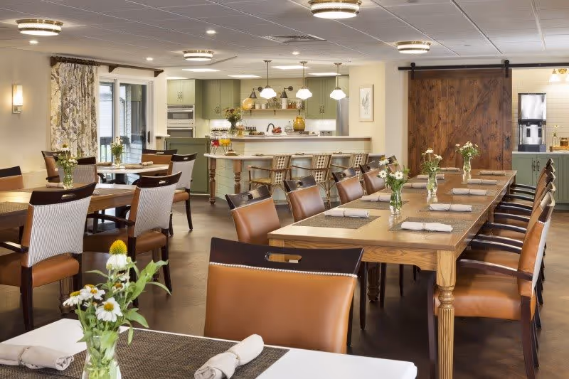 A bright and spacious dining room with multiple wooden tables and chairs arranged neatly. Each table has a small vase with white flowers and rolled napkins placed on woven placemats. In the background, there is a kitchen area with green cabinetry, a countertop with bar stools, and pendant lights hanging from the ceiling. A large wooden sliding barn door is visible on the right side of the kitchen area.