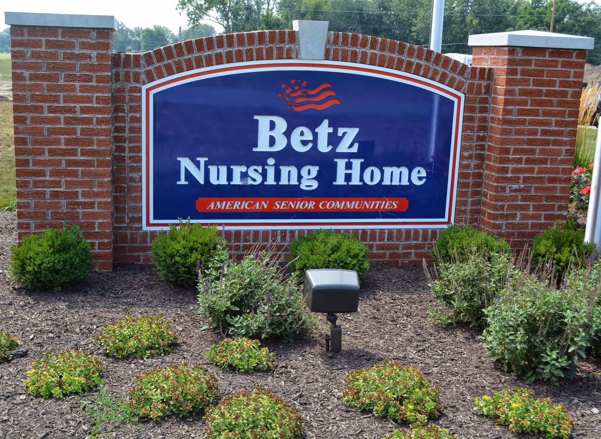 A large brick sign for Betz Nursing Home with a blue background and white text, featuring a small American flag graphic and the words 'American Senior Communities' below. The sign is surrounded by landscaping with small bushes and plants.