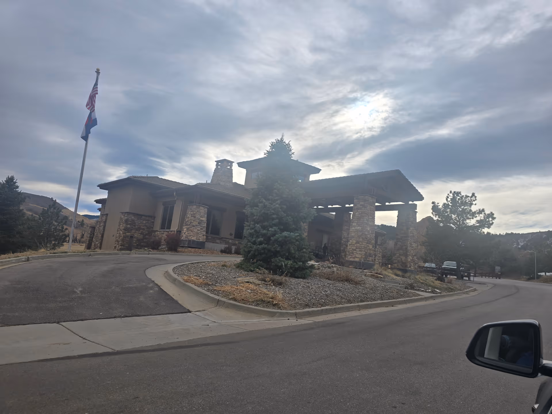 Exterior view of a single-story building with stone pillars and a covered entrance, surrounded by trees and a flagpole with two flags. The sky is cloudy with the sun partially visible behind the clouds.