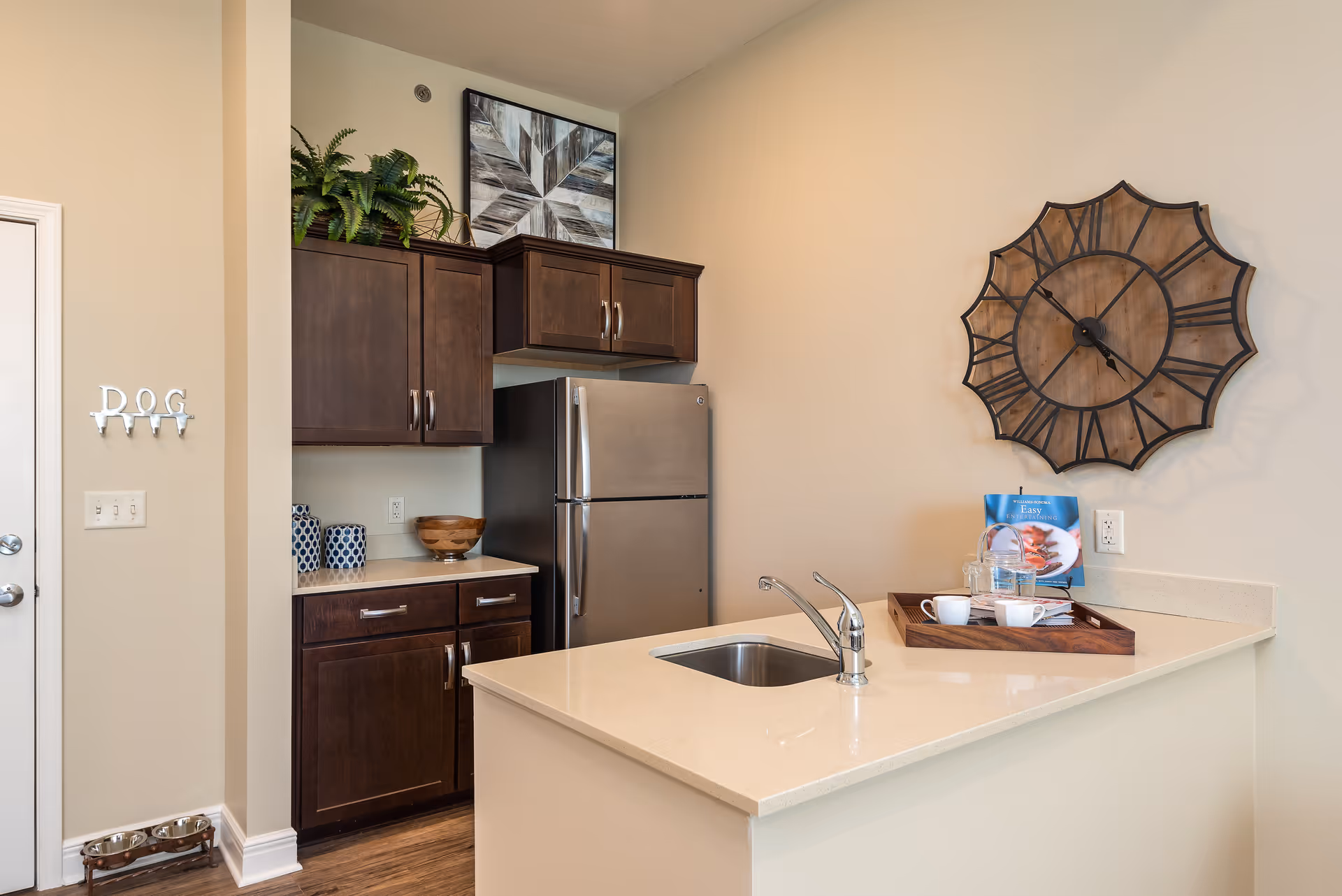 A modern kitchen area with dark wood cabinets, a stainless steel refrigerator, and a light-colored countertop with a built-in sink. On the countertop is a wooden tray holding a glass pitcher, two white cups, and a cookbook. A large decorative wall clock with Roman numerals hangs on the wall above the countertop. There is a small plant and decorative items on top of the cabinets. On the left wall, there is a metal hook rack labeled 'DOG' above two pet bowls on the floor.