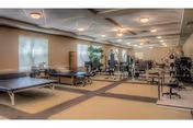 A spacious physical therapy or rehabilitation room with multiple exercise tables and various exercise equipment arranged neatly. The room has large windows with blinds, beige walls, and a patterned carpet floor. Ceiling lights provide ample illumination.