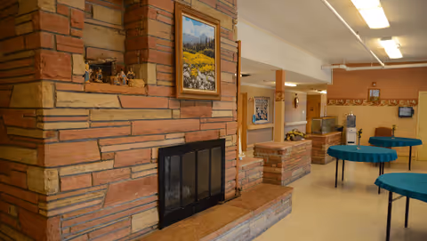 Interior view of a senior living facility common area featuring a large stone fireplace with a framed landscape painting above it. The room has several round tables covered with teal tablecloths, a water dispenser, and a fish tank. The walls are decorated with a floral border and various pictures, and the ceiling has fluorescent lighting.