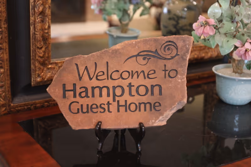 A decorative stone sign on a stand reading "Welcome to Hampton Guest Home" displayed on a table with potted plants and a mirror behind it.