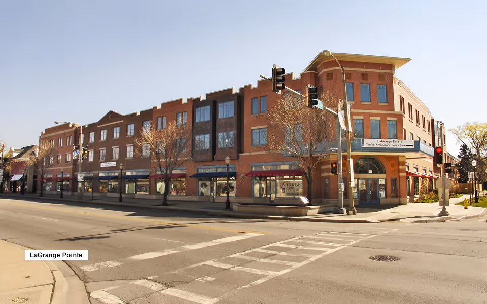 Exterior view of a three-story brick building on a street corner with traffic lights and crosswalks. The building has multiple windows and storefronts on the ground floor. A sign on the corner reads 'LaGrange Pointe Senior Residence Now Open.' Leafless trees line the sidewalk in front of the building.