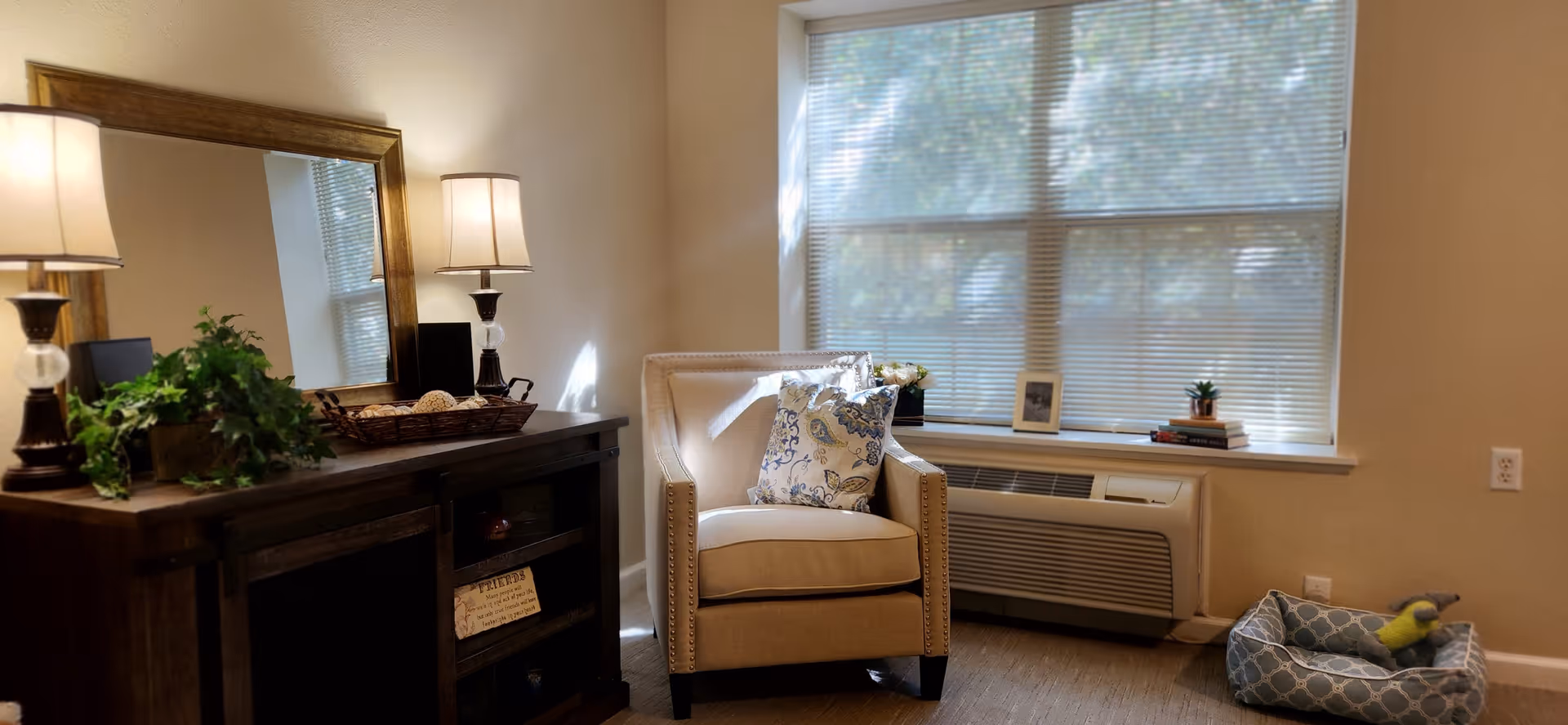 A cozy living room area with a beige armchair adorned with two decorative pillows, positioned near a window with closed blinds. To the left, there is a dark wooden cabinet with a large mirror above it, two table lamps, and a green potted plant. On the right side, a pet bed with a toy is placed on the floor next to the wall.