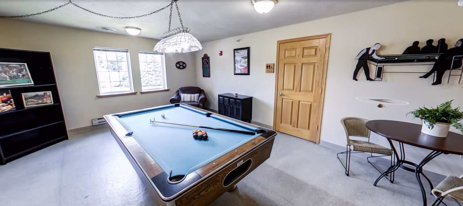 A recreational room with a pool table in the center, two pool cues resting on the table, a rack of billiard balls, a round table with two chairs and a potted plant, a wooden door, a cushioned armchair, a black cabinet, a bookshelf with framed pictures, two windows letting in natural light, and a wall decoration depicting two people playing pool.