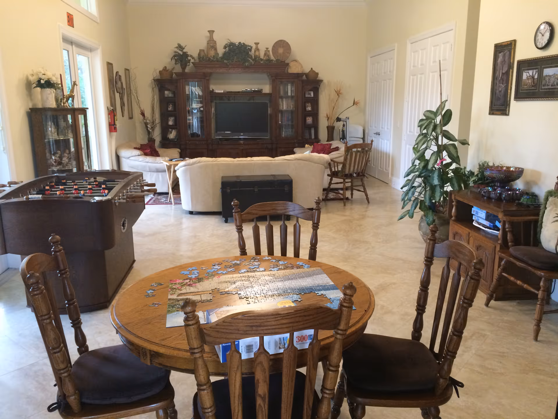 Spacious common living room with a round table holding a jigsaw puzzle in the foreground, a foosball table to the left, sofas and an entertainment center in the background.