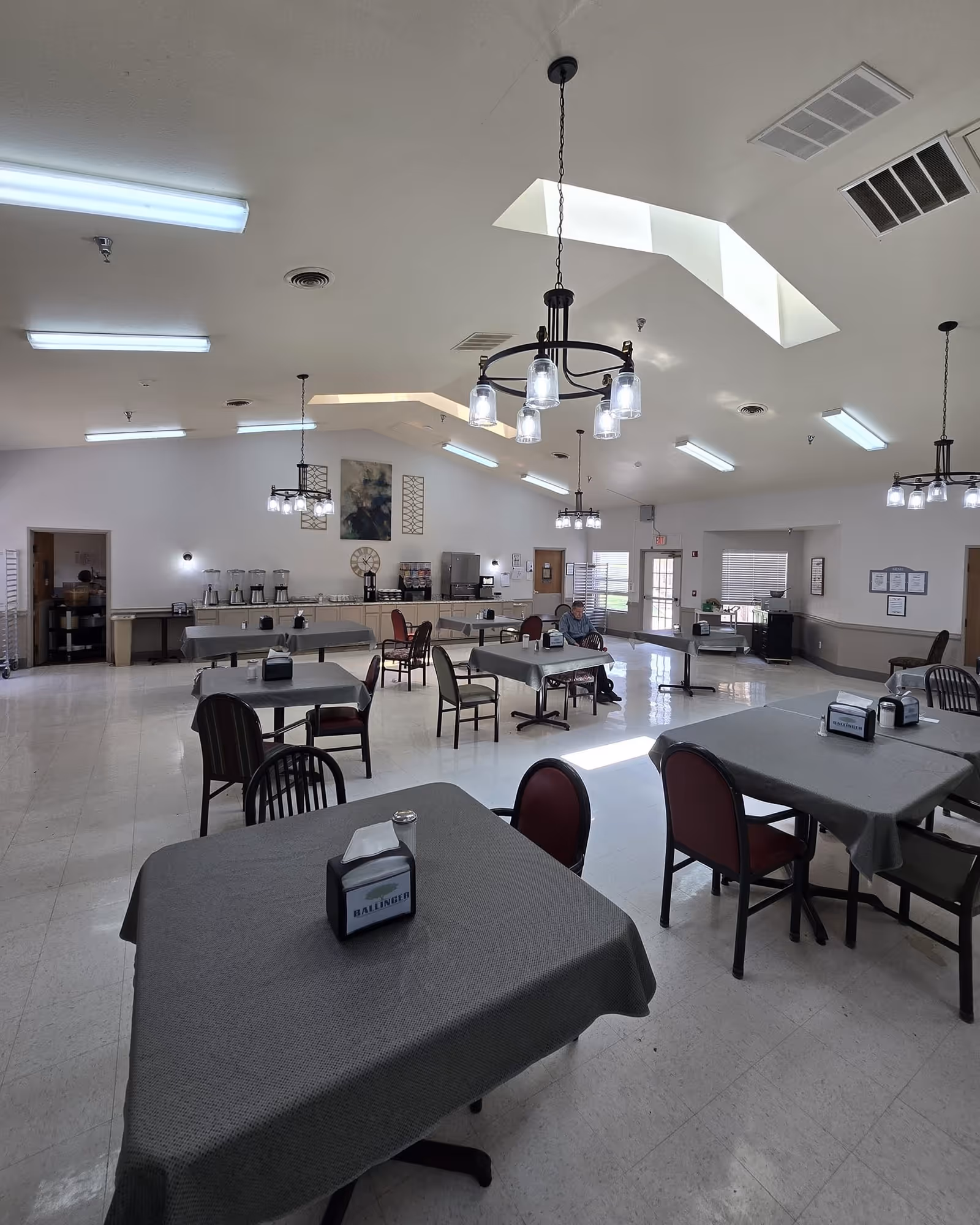 A spacious dining room with multiple tables covered in gray tablecloths, each with a napkin holder labeled 'Ballinger'. The room has several hanging light fixtures, a high ceiling with skylights, and a beverage station along the far wall. A single person is seated at one of the tables near the back.
