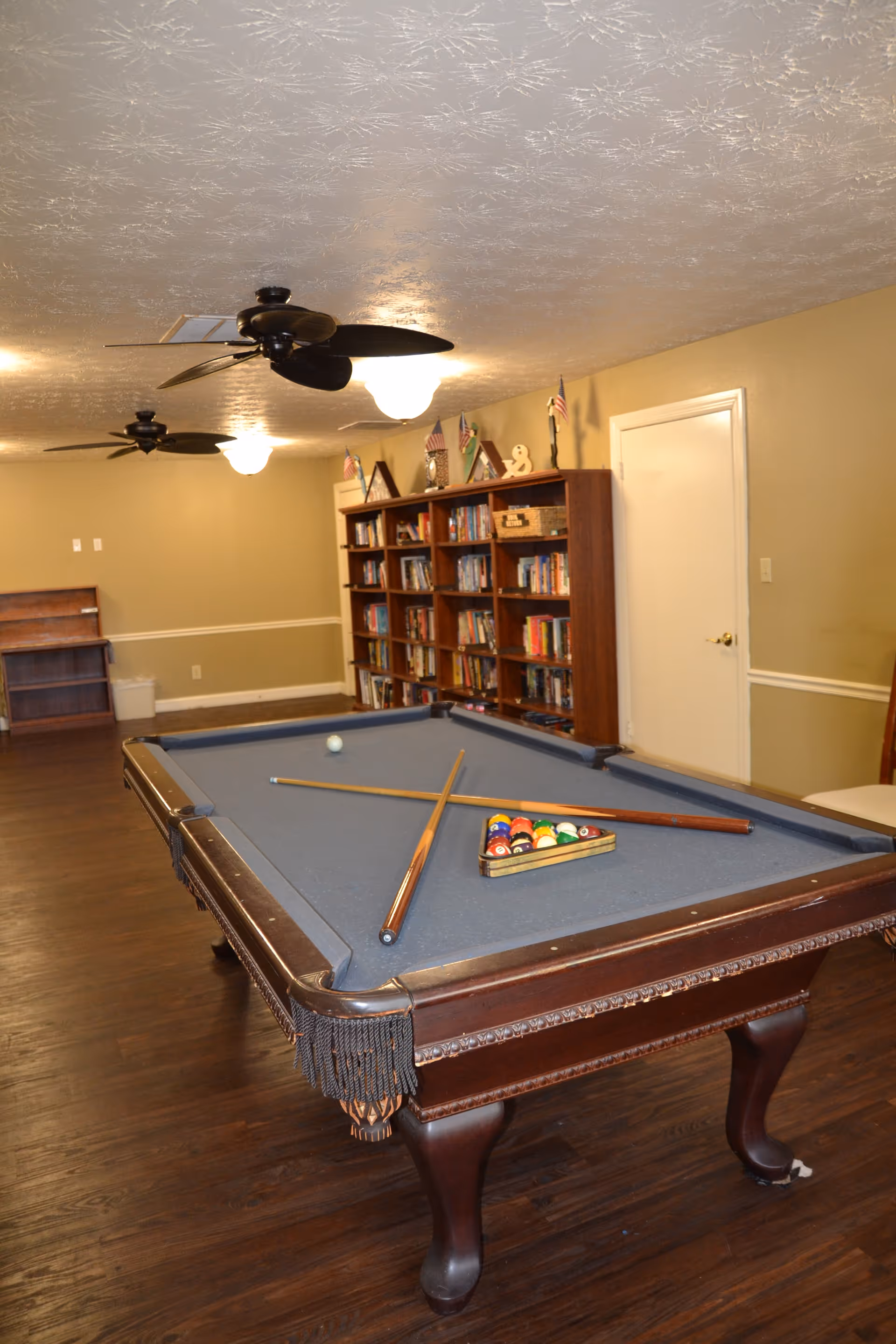 A room with a pool table in the center, two pool cues resting on the table, and a rack of billiard balls arranged in a triangle. The room has wooden flooring, beige walls, a bookshelf filled with books and small American flags on top, ceiling fans, and overhead lights.