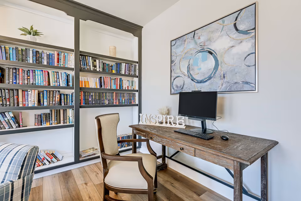 A cozy reading and work area with a wooden desk holding a computer monitor, keyboard, and mouse. A decorative sign spelling 'INSPIRE' is placed on the desk. Behind the desk is a large abstract painting with circular patterns. To the left, there is a built-in bookshelf filled with books and a small potted plant on top. A cushioned wooden chair is positioned in front of the desk, and part of a striped armchair is visible in the foreground. The room has light-colored walls and wood flooring.