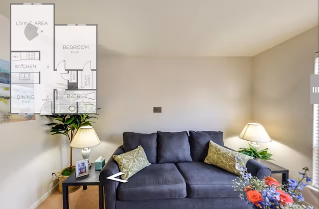 A bright living room with a gray sofa topped with patterned pillows, side tables with lamps, plants and a bouquet, and a small floor-plan overlay in the top-left corner.