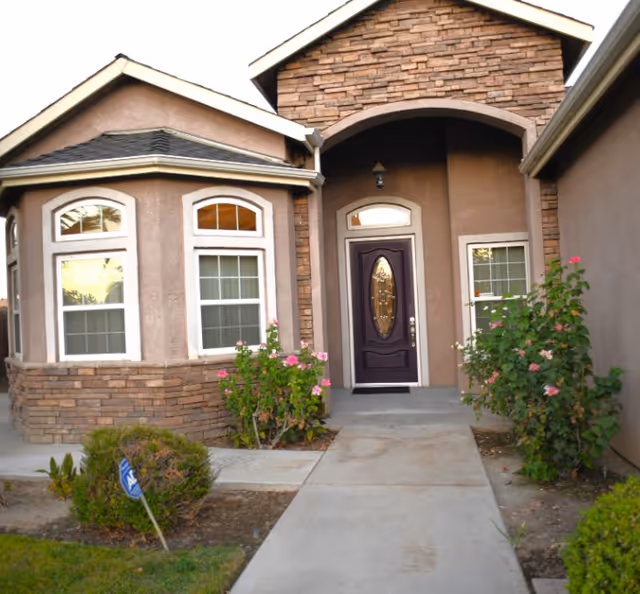 Front entrance of a residential building with a dark wooden door featuring an oval glass panel. The entrance is framed by stone and stucco walls, with two windows on either side and flowering bushes along the walkway leading to the door.