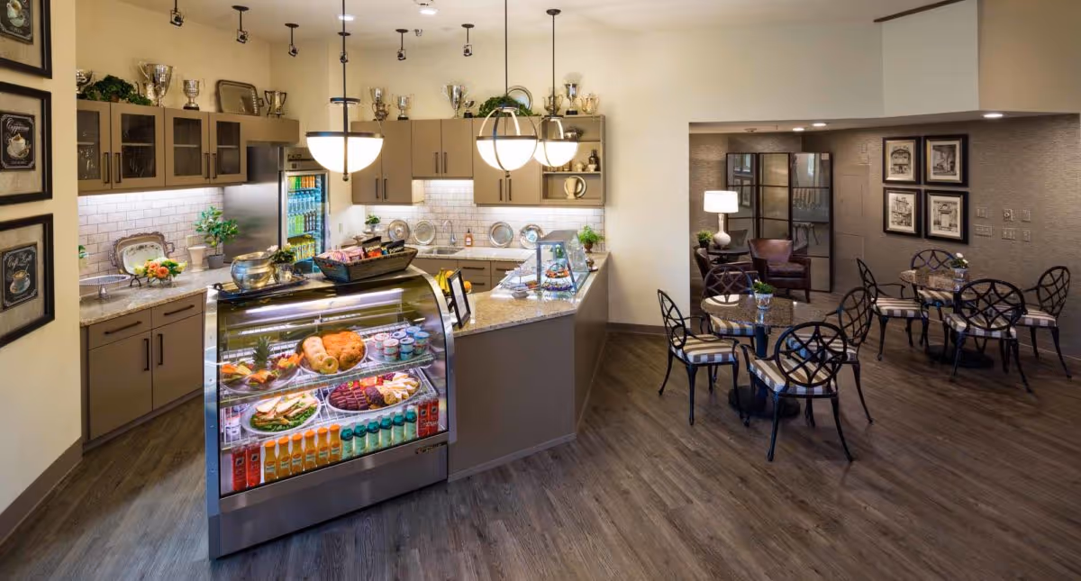 Interior view of a modern assisted living community dining and snack area featuring a glass display case with assorted food and beverages, a countertop with decorative items, hanging pendant lights, and several round tables with chairs arranged for seating. The space has wood flooring, framed artwork on the walls, and a cozy seating area with a lamp and armchair in the background.