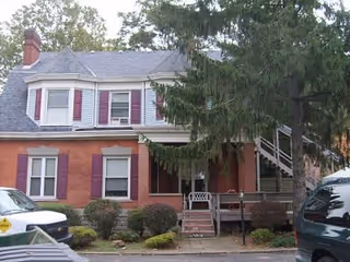 Exterior view of a two-story brick building with white trim and red shutters, surrounded by bushes and trees. There is a porch with a bench and stairs leading up to it, and a large evergreen tree partially obscures the right side of the building. Several vehicles are parked in front of the building.