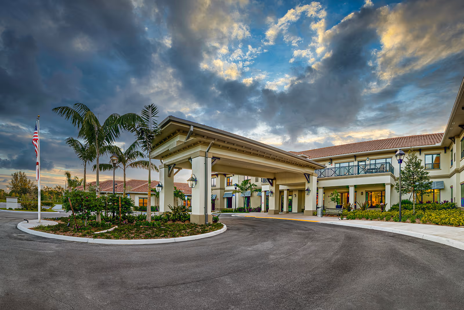 Front entrance and porte-cochere of a senior living building with landscaping, palm trees, and an American flag beneath a dramatic cloudy sky.