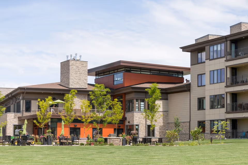 Exterior front view of a modern senior living building with balconies, patio seating, and a manicured lawn.