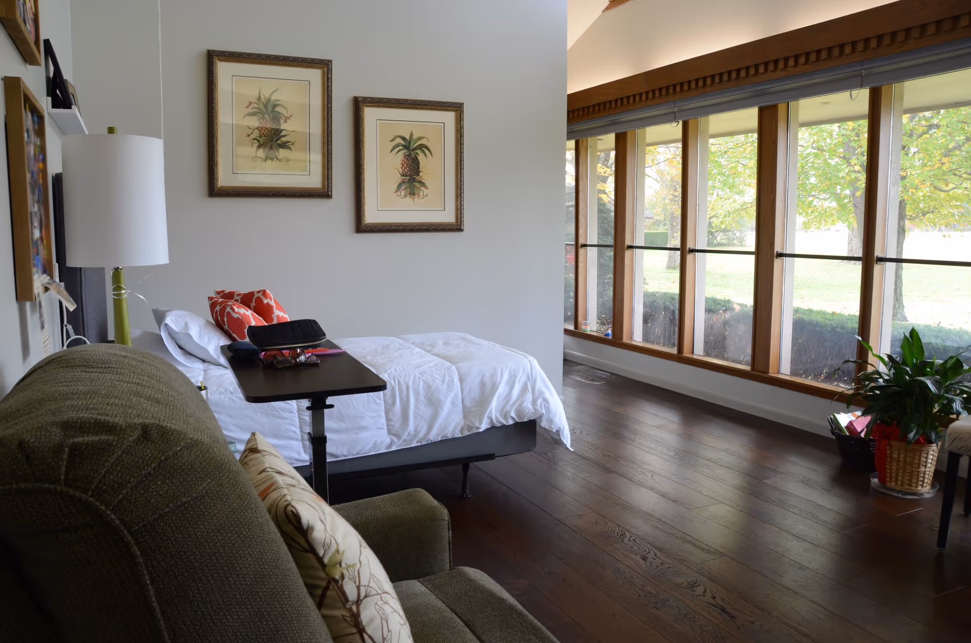Sunlit bedroom with a bed, armchair, bedside table, and large floor-to-ceiling windows overlooking greenery.