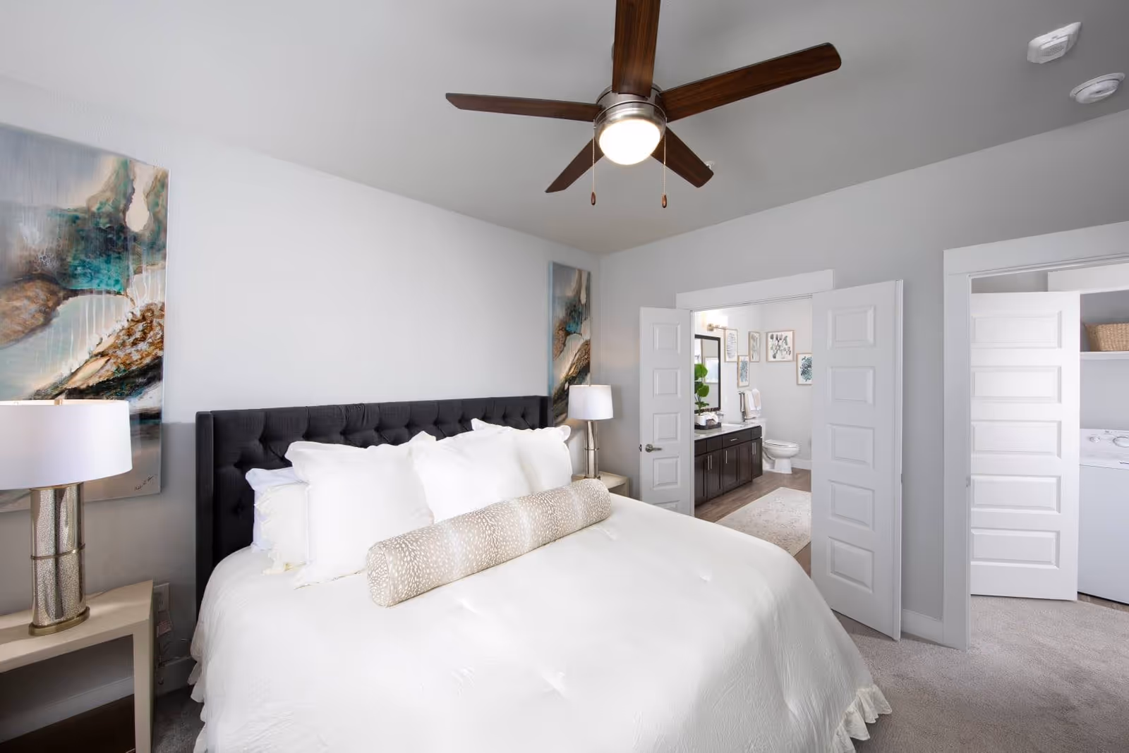 Bright bedroom with a made bed, ceiling fan, nightstands and open doors revealing a bathroom and laundry area.