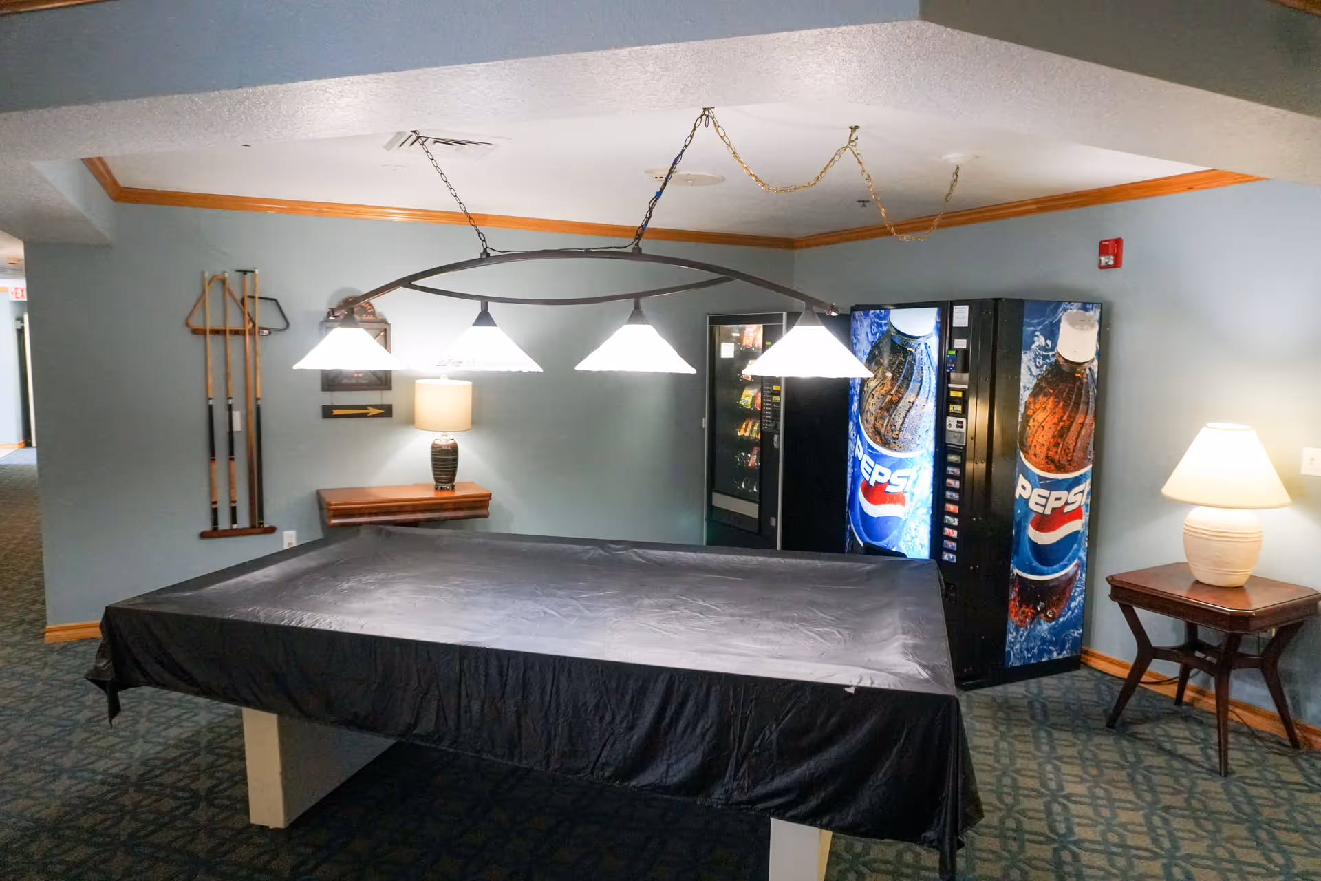 Indoor recreational area with a covered pool table under four hanging lights, a wall-mounted rack holding pool cues, two Pepsi vending machines, and two side tables each with a lamp, all set against light blue walls with wood trim.