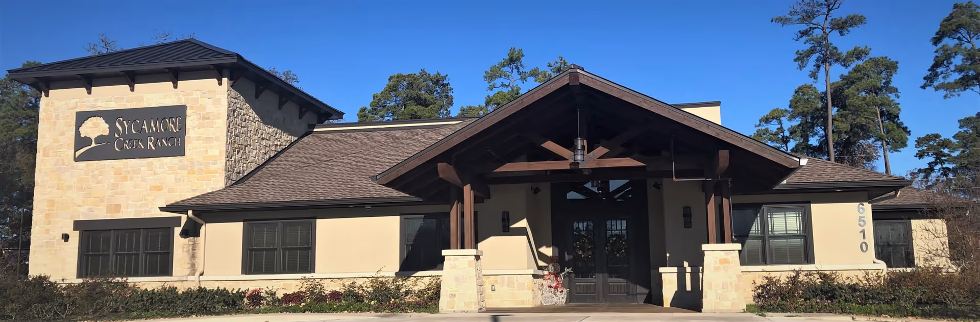 Front exterior view of Sycamore Creek Ranch at Cypresswood building with a stone and beige facade, dark roof, large wooden entrance structure, and surrounding greenery under a clear blue sky.