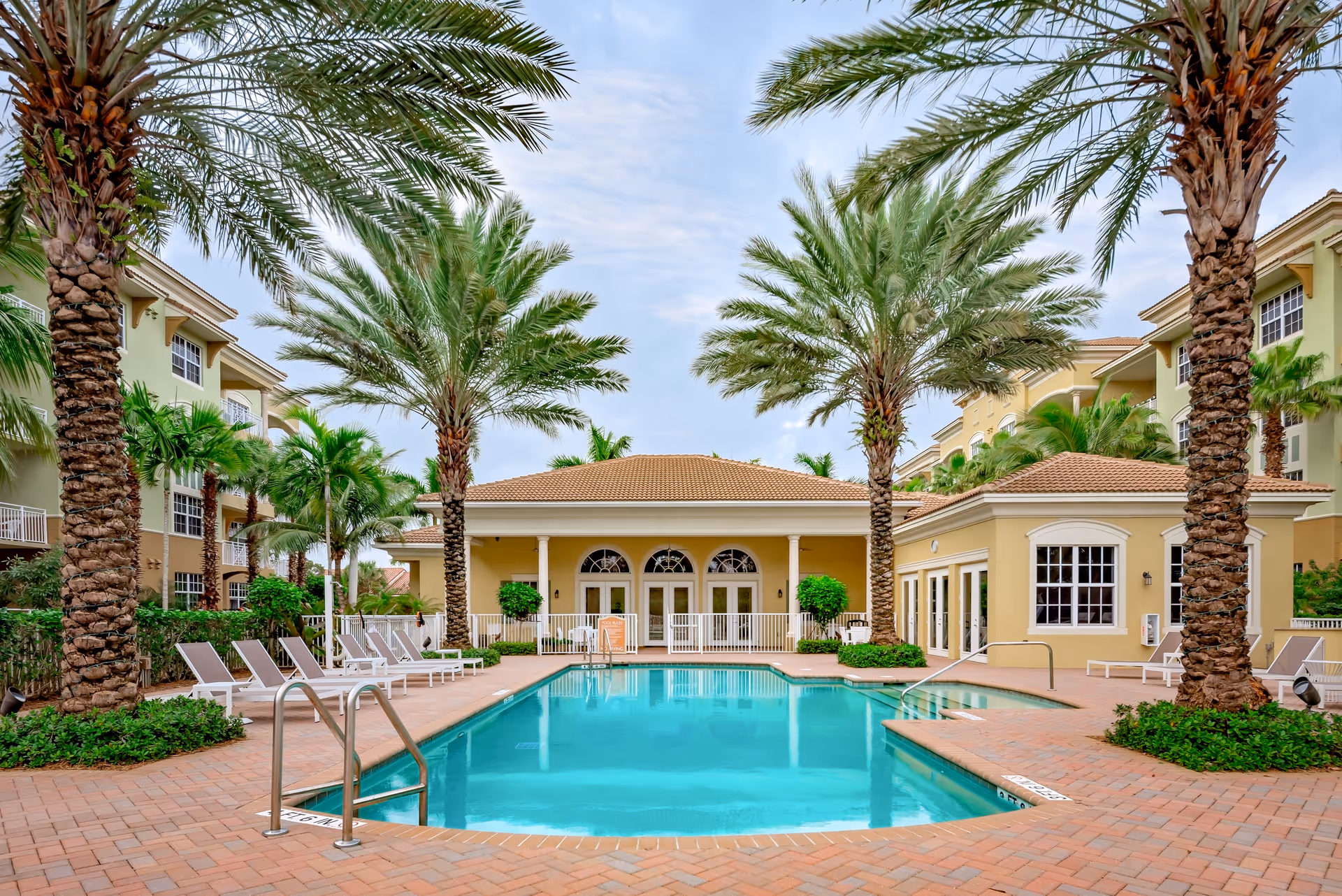 Outdoor swimming pool area at Riverwalk Pointe facility, surrounded by palm trees and lounge chairs, with a beige building in the background under a partly cloudy sky.