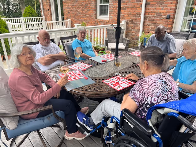 Six elderly individuals sitting around a round outdoor table on a patio, playing a game of bingo. The table has bingo cards and glasses with drinks. The setting is outside a brick building with white railings and windows.