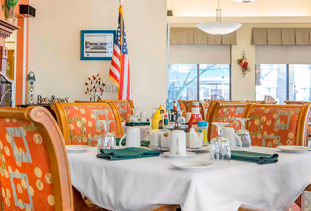A dining table set with white tablecloth, green napkins, glassware, plates, and condiments including ketchup and mustard in a bright dining room with patterned orange chairs. An American flag and framed picture are visible on the wall in the background.