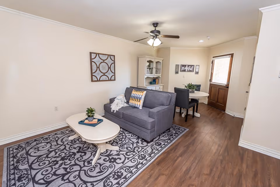 A cozy living room area with a gray couch adorned with a throw blanket and a decorative pillow, a white oval coffee table with a small plant and books on it, a patterned area rug underneath, and a white cabinet with shelves in the background. There is a small dining area with a round white table and two black chairs near a wooden door with a window. The walls are light-colored, and the floor is wooden with a ceiling fan overhead.
