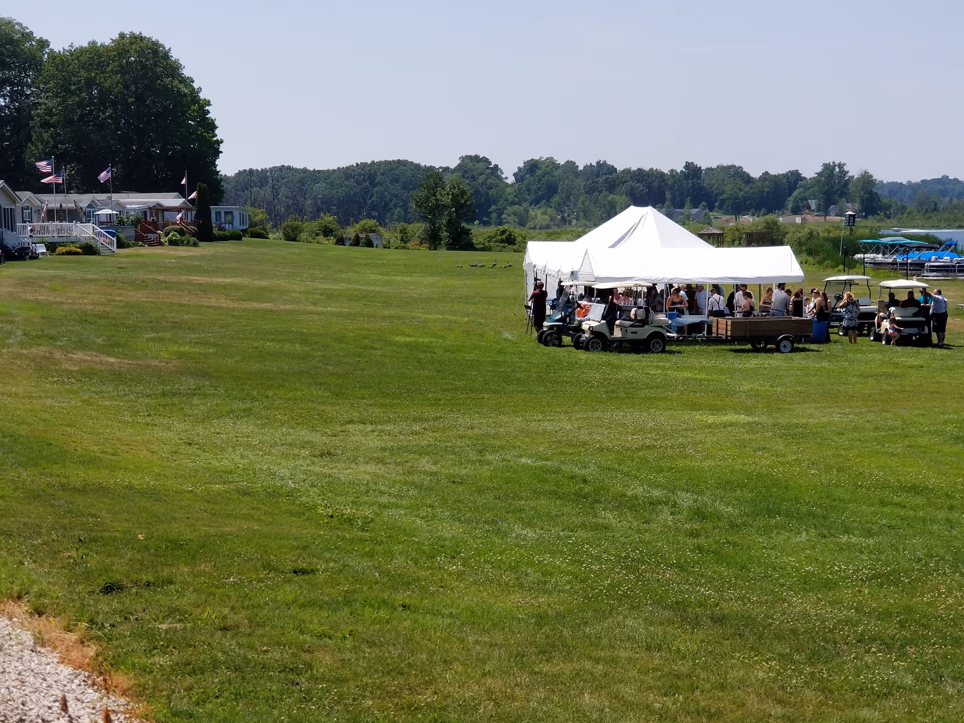 A large grassy outdoor area with a white event tent set up where a group of people are gathered. Several golf carts are parked near the tent. In the background, there are trees, a few houses with American flags, and a lake with boats docked.