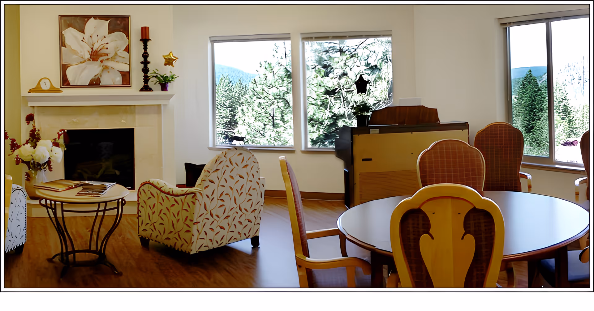 A cozy living room area with a fireplace, floral armchairs, a round wooden table with chairs, and large windows showing a view of trees and mountains outside. The room is decorated with a large flower painting, a candle holder, and a small clock on the fireplace mantel.