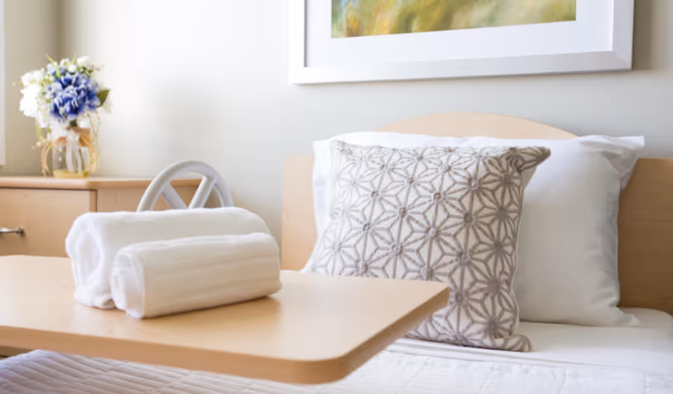 A tidy bedroom in a senior living facility with a tray table, towels, and floral decor.