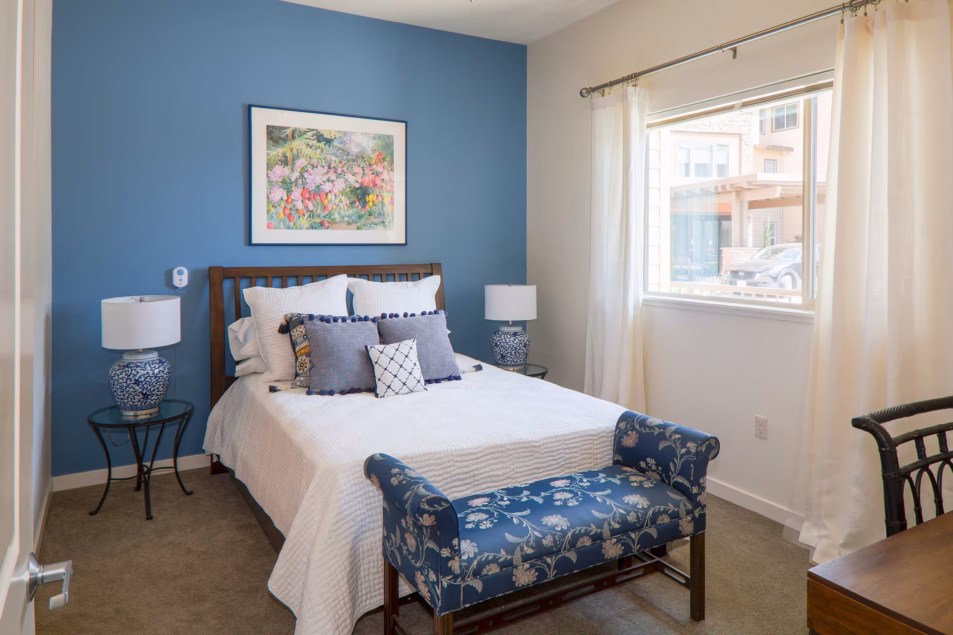 A cozy bedroom with a wooden bed frame against a blue accent wall. The bed is neatly made with white bedding and several decorative pillows in shades of blue and white. On each side of the bed are matching blue and white ceramic lamps on glass-top side tables. A floral painting hangs above the bed. A window with sheer white curtains lets in natural light, and a blue floral upholstered bench sits at the foot of the bed. A wooden chair and desk are partially visible on the right side of the room.
