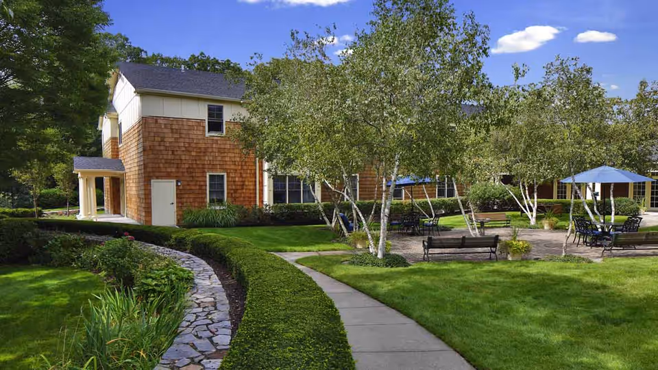A landscaped courtyard with paved walkways, benches, tables with umbrellas, and a shingled two-story building in the background.