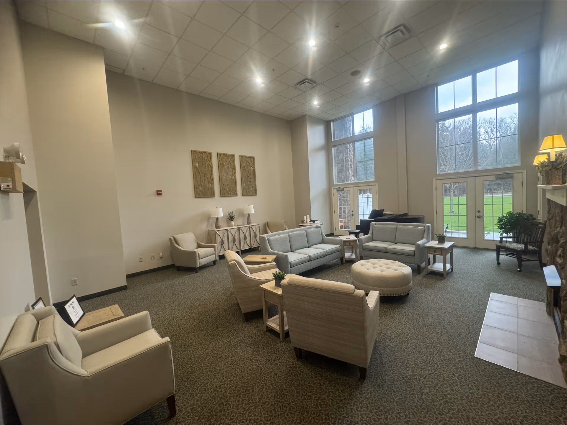 Bright, spacious common lounge area with multiple sofas, armchairs, a round ottoman, side tables and tall windows overlooking a green lawn.