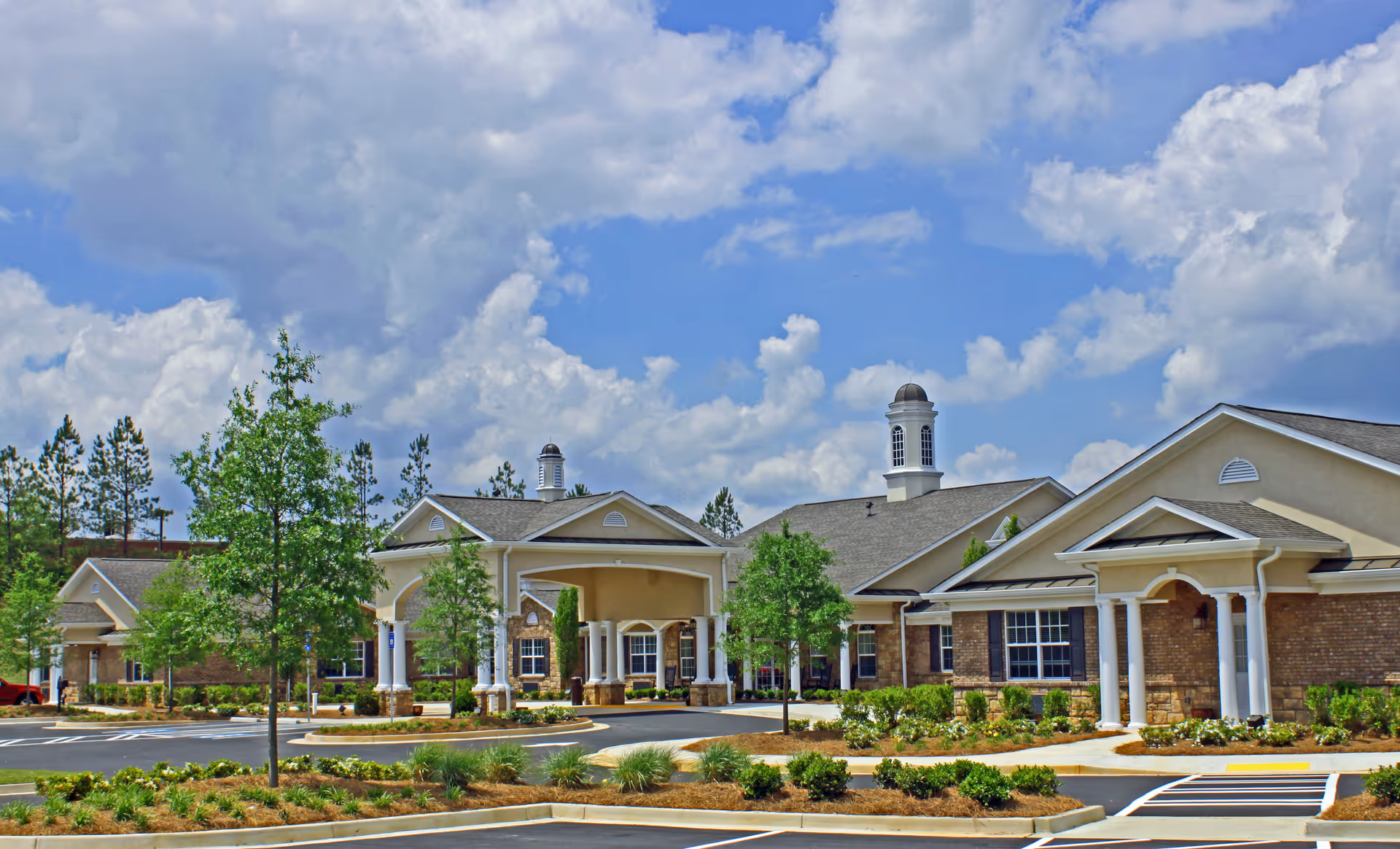 Exterior view of Benton House of Staley Hills, a single-story senior living facility with beige walls, brick accents, white columns, and cupolas on the roof. The building is surrounded by landscaped greenery and a parking lot under a partly cloudy blue sky.