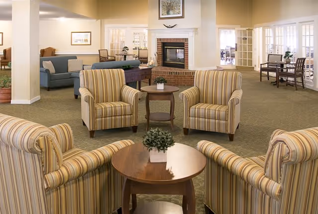 A spacious senior living community lounge area with four striped armchairs arranged around a round wooden table with a small potted plant. In the background, there is a brick fireplace with a framed picture above it, additional seating including a blue sofa, and glass-paned doors leading to other rooms.