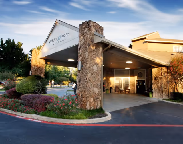 Entrance of West Fork Village senior living facility with a covered drop-off area supported by stone pillars, surrounded by landscaped bushes and flowers under a partly cloudy sky.