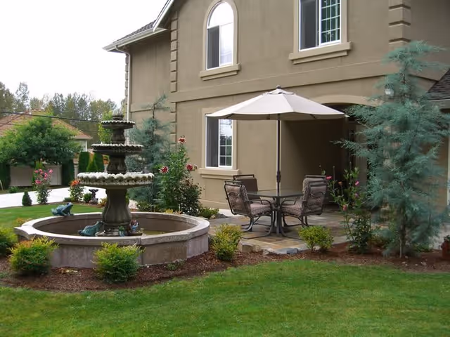 Outdoor patio area with a round table and four chairs under a large beige umbrella, surrounded by greenery and flowering plants. A multi-tiered stone fountain is in the foreground, and the exterior of a beige building with windows is visible in the background.