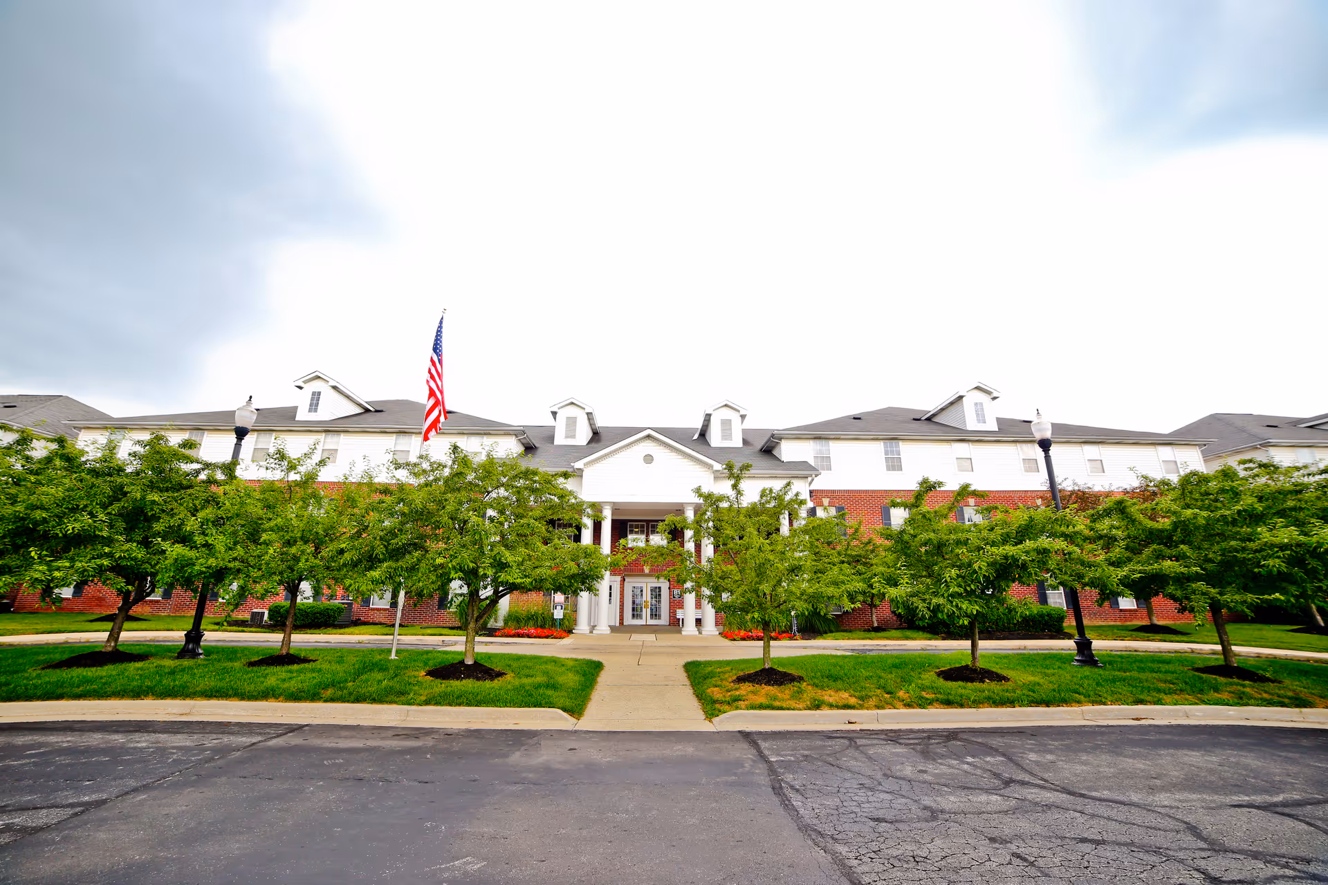 Front exterior view of a large senior living facility named Benjamin Court with a brick and white facade, multiple windows, a central entrance with columns, an American flag, and neatly trimmed trees and grass in front.