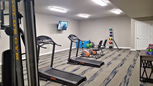 Indoor fitness room with two treadmills, two recumbent exercise bikes, a rack of colorful medicine balls, a stack of blue exercise mats, and a wall-mounted TV. The room has beige walls and patterned carpet flooring.