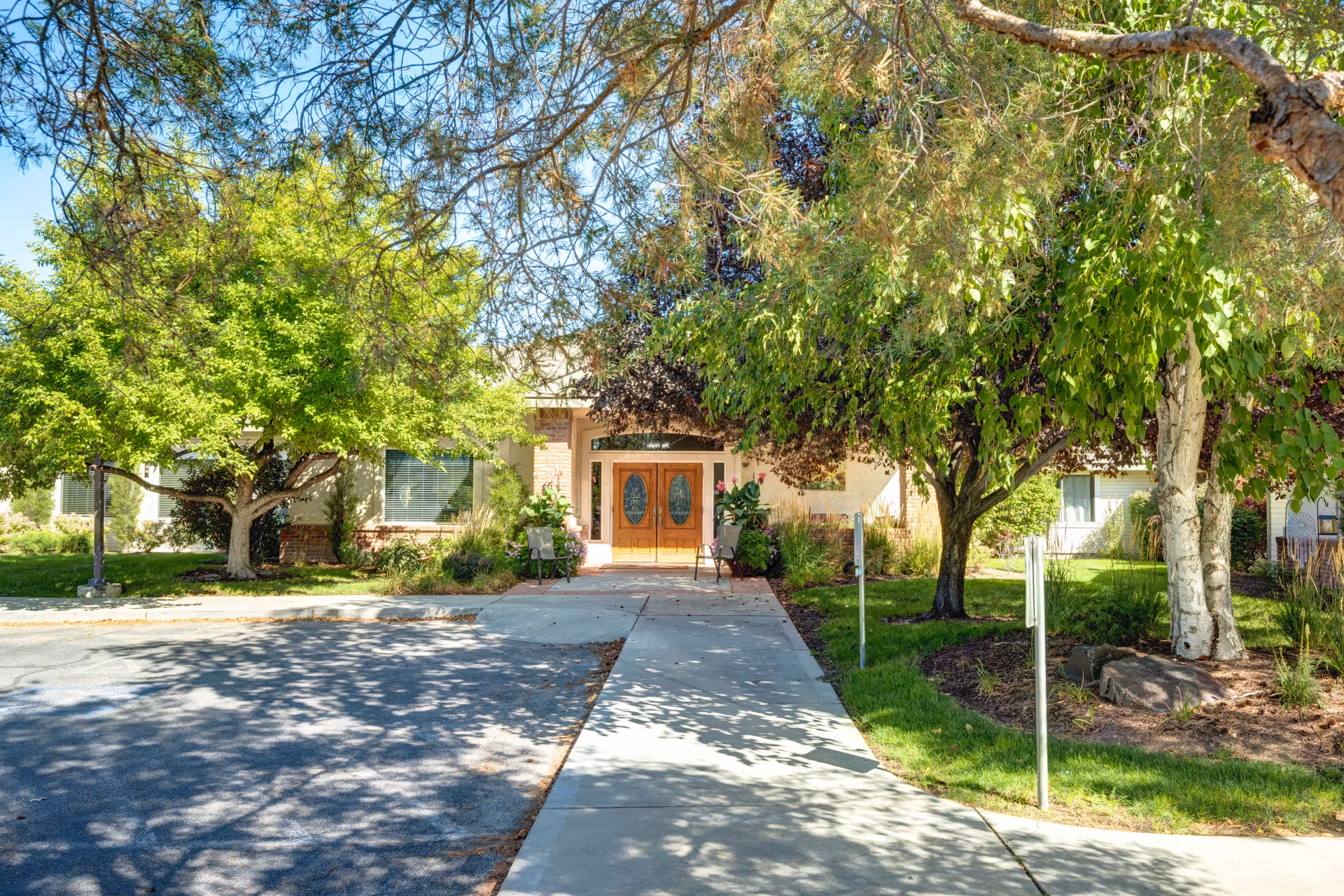 Front entrance of Overland Court Senior Living facility with a concrete walkway leading to double wooden doors with oval glass panels. The entrance is flanked by two chairs and surrounded by lush green trees and landscaping under a clear blue sky.