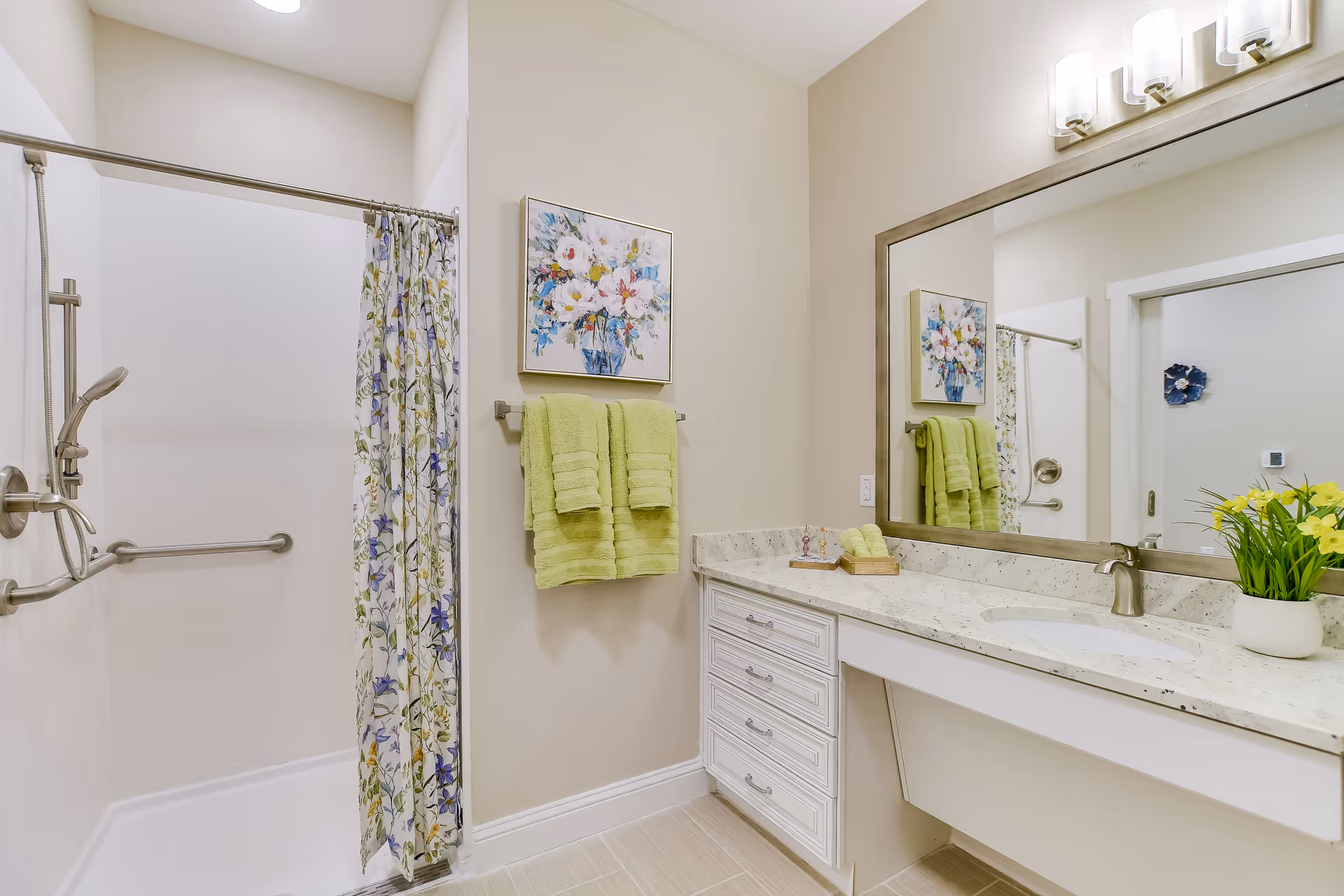 A bright and clean bathroom featuring a walk-in shower with a floral shower curtain, a wall-mounted grab bar, and a handheld showerhead. Next to the shower, two green towels hang on a towel rack beneath a framed floral painting. A large mirror above a white vanity with a marble countertop reflects part of the room. On the countertop, there is a small tray with soap and a potted plant with yellow flowers. The walls are painted a soft beige color.