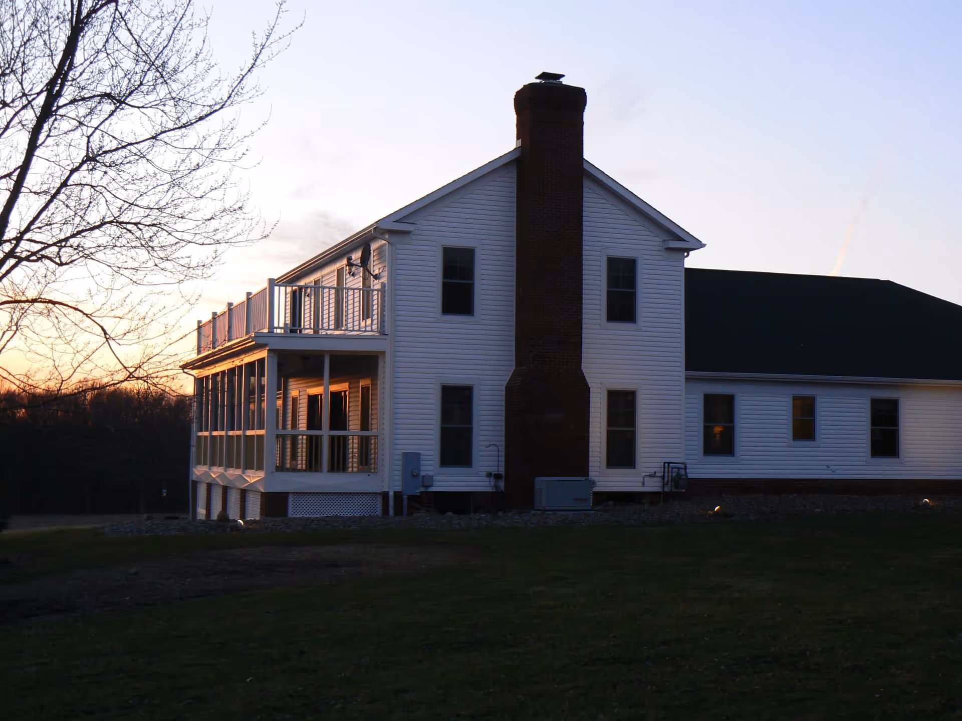 A white two-story building with a large brick chimney, multiple windows, and a balcony with railings, set against a sunset sky with bare tree branches visible on the left side and a grassy lawn in the foreground.
