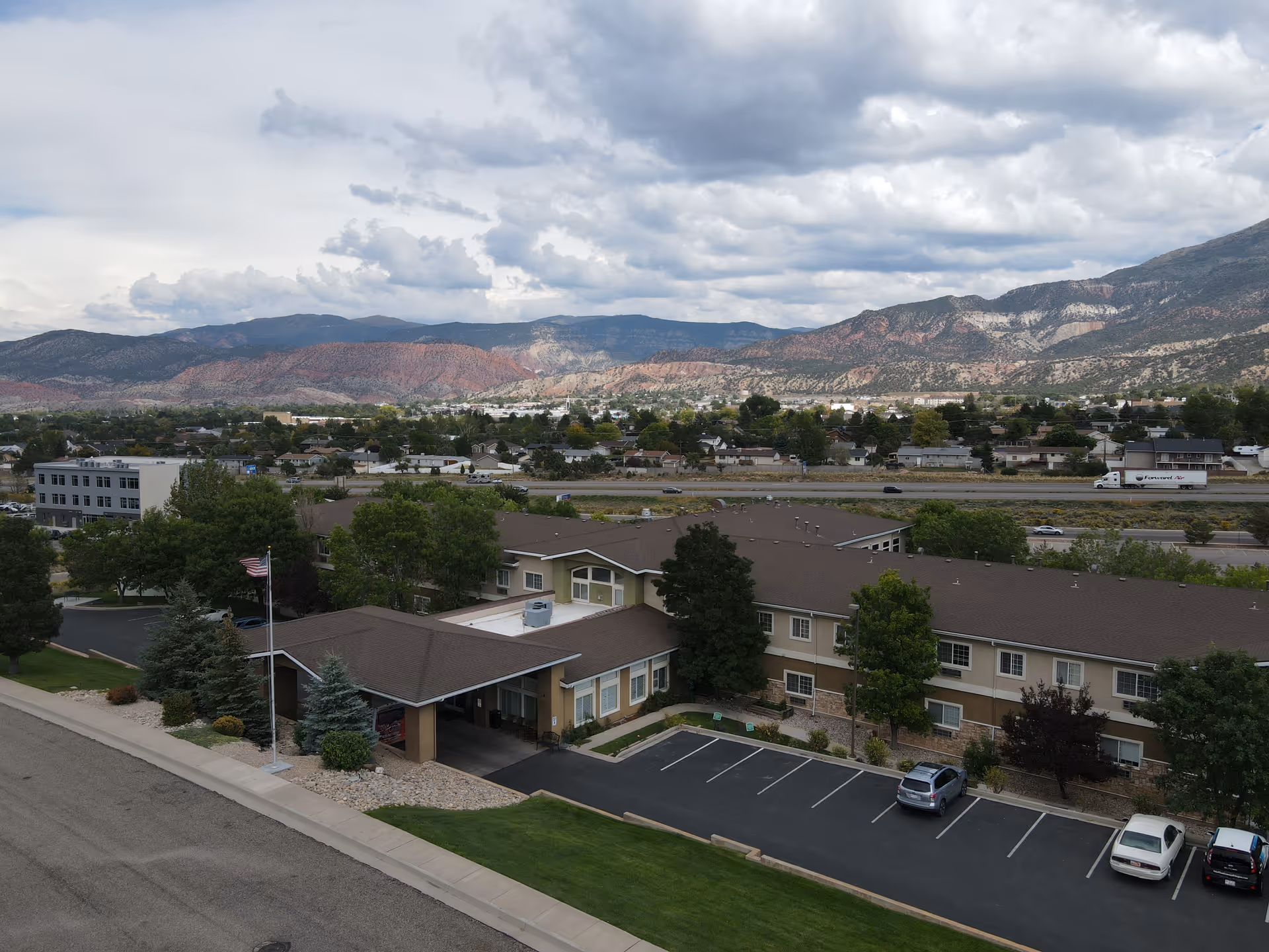 Aerial view of Our House Assisted Living of Cedar City, showing a two-story building with a brown roof, surrounded by trees and parking spaces. In the background, there is a residential neighborhood and mountains under a cloudy sky.