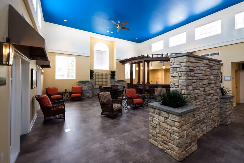 A spacious and well-lit common area with a high blue ceiling and ceiling fan. The room features several comfortable chairs with red and brown cushions arranged around small tables. There is a stone partition with built-in planters and a wooden pergola structure over a counter or bar area. Large windows allow natural light to fill the space.