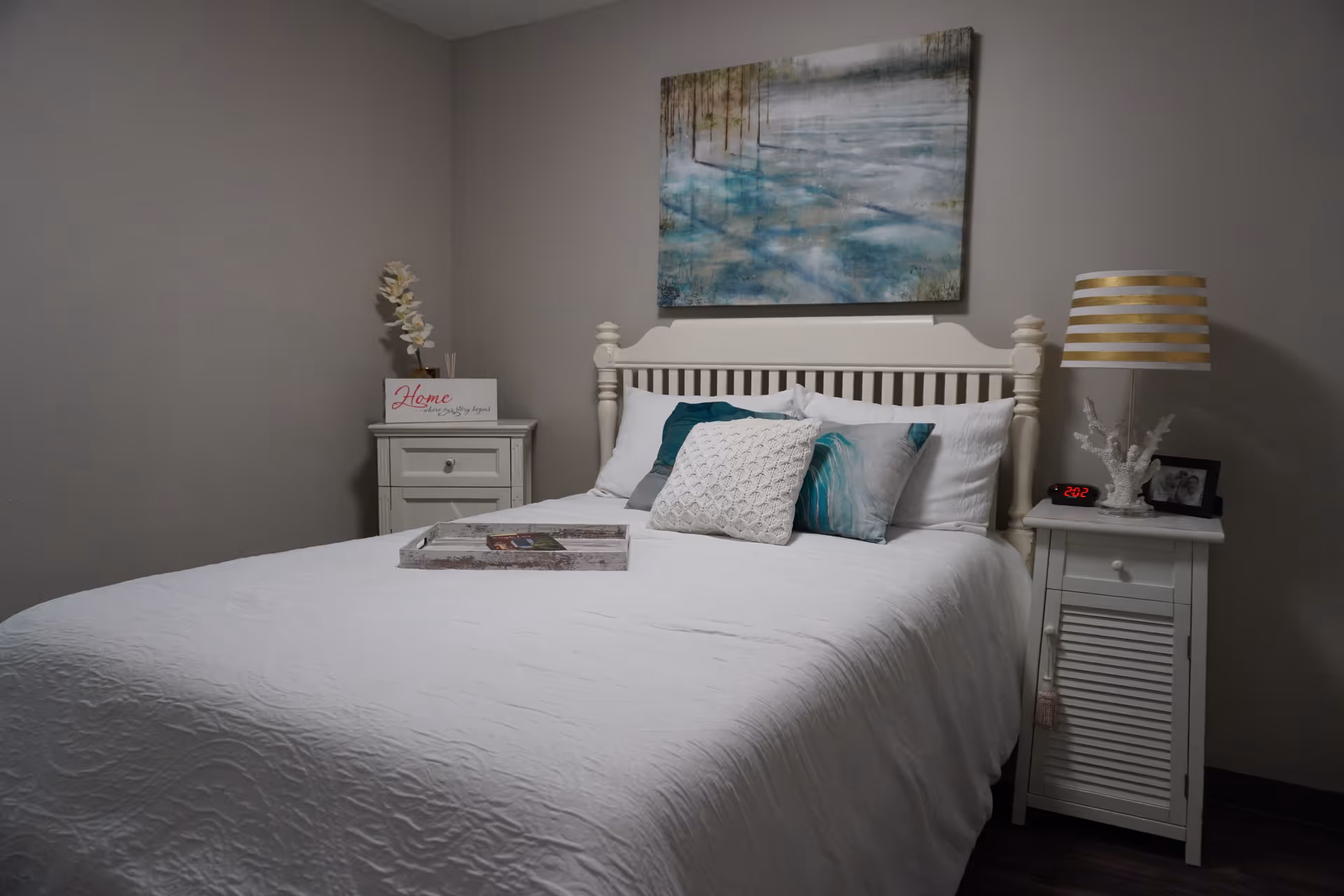 A neatly made bed with white bedding and multiple pillows in a bedroom. There are two white nightstands on either side of the bed. The left nightstand has a decorative flower and a sign that reads 'Home where your story begins.' The right nightstand has a lamp with a gold and white striped shade, a digital clock showing 2:02, a white coral decoration, and a framed photo. Above the bed hangs a large painting with abstract blue and green tones.