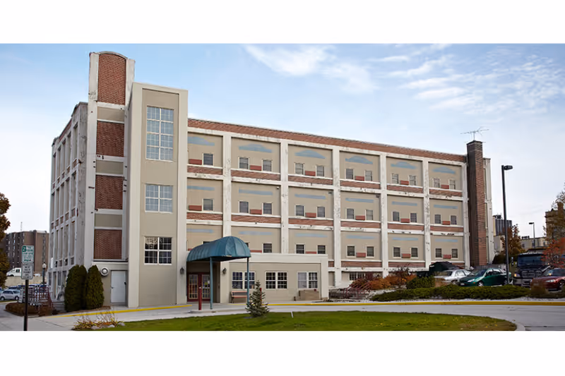 Four-story beige and brick assisted living building with a covered entrance and parking lot in front.