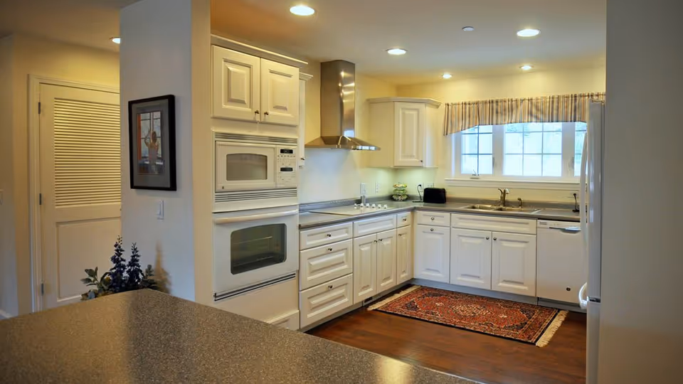 A bright kitchen with white cabinets, a built-in microwave and oven, a stainless steel range hood, a double sink under a window with striped valance, a dishwasher, and a refrigerator. There is a patterned rug on the wooden floor and a countertop in the foreground.