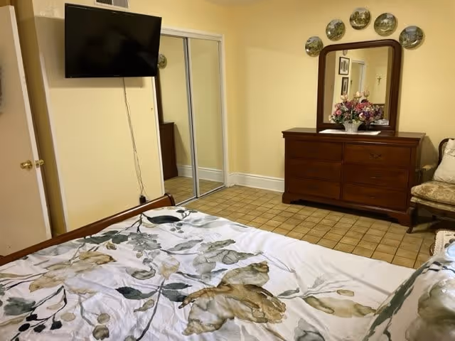 A bedroom with a bed covered in a floral patterned comforter in the foreground. The room has a wooden dresser with a mirror and a vase of flowers on top. Above the dresser, decorative plates are arranged on the wall. There is a chair with a cushion next to the dresser. A flat-screen TV is mounted on the wall opposite the bed, and a closet with sliding mirrored doors is visible. The floor is tiled and the walls are painted light yellow.