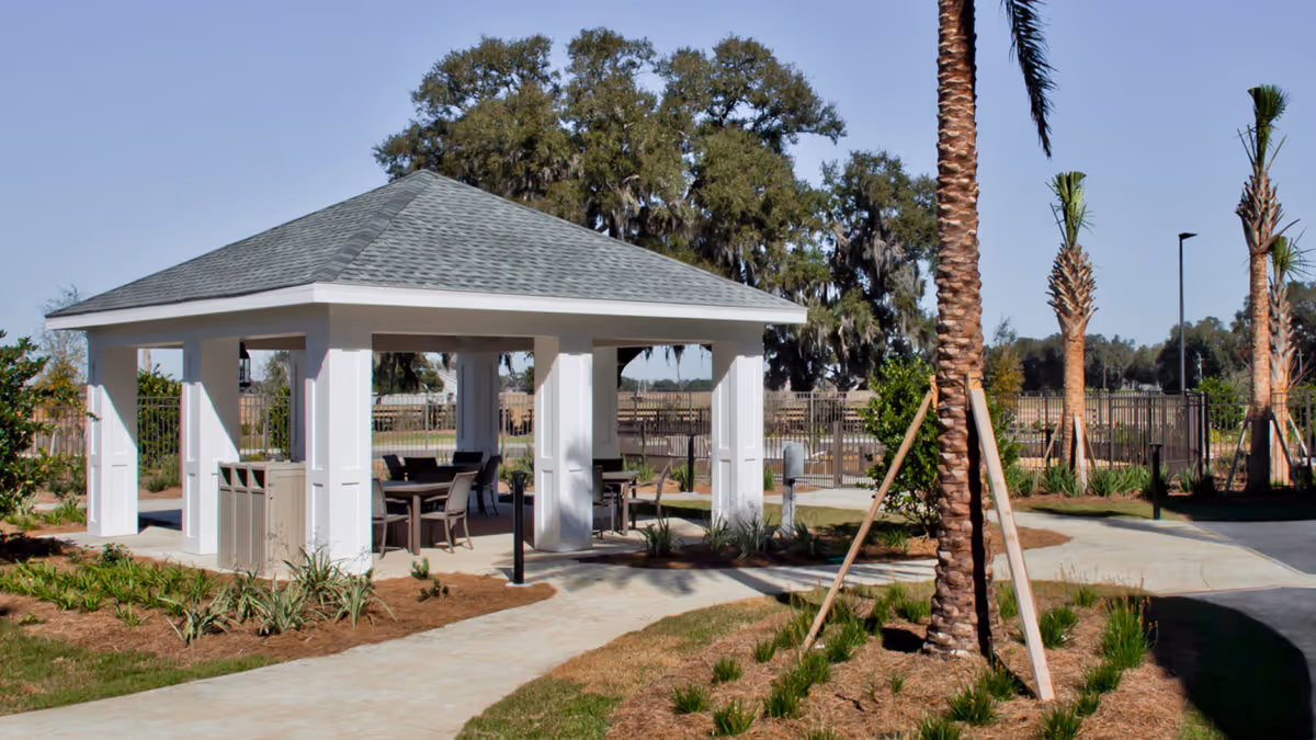 A white outdoor pavilion with tables and chairs surrounded by palm trees and landscaped walkways.